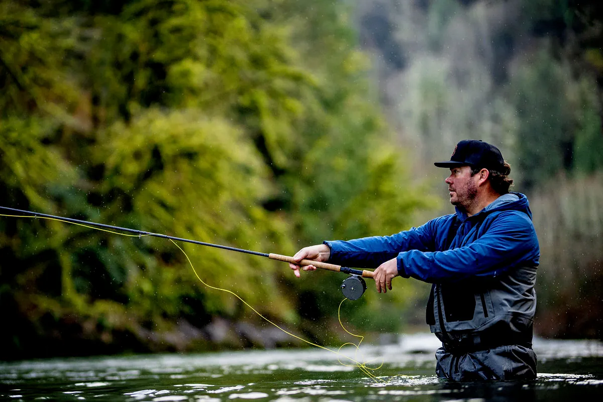 Chris O'Donnell Spey casting a two-handed fly rod while wading a rain-dappled Oregon coastal steelhead river lined with green timber.