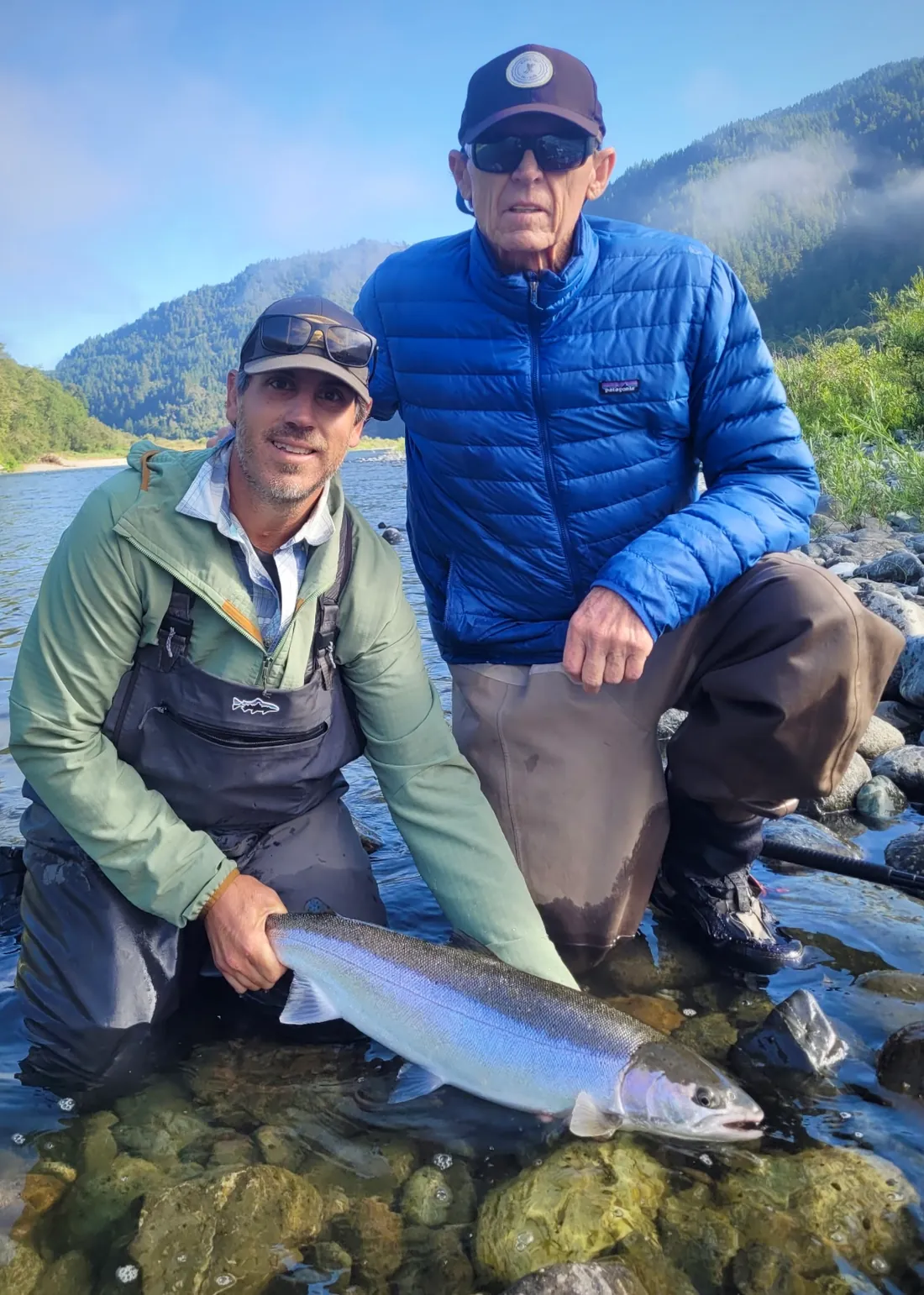 Jason Hartwick, in a green jacket and waders, kneeling in clear water over mossy cobbles beside an angler in a blue down jacket, cradling a bright wild steelhead in a forested Northern California river canyon.