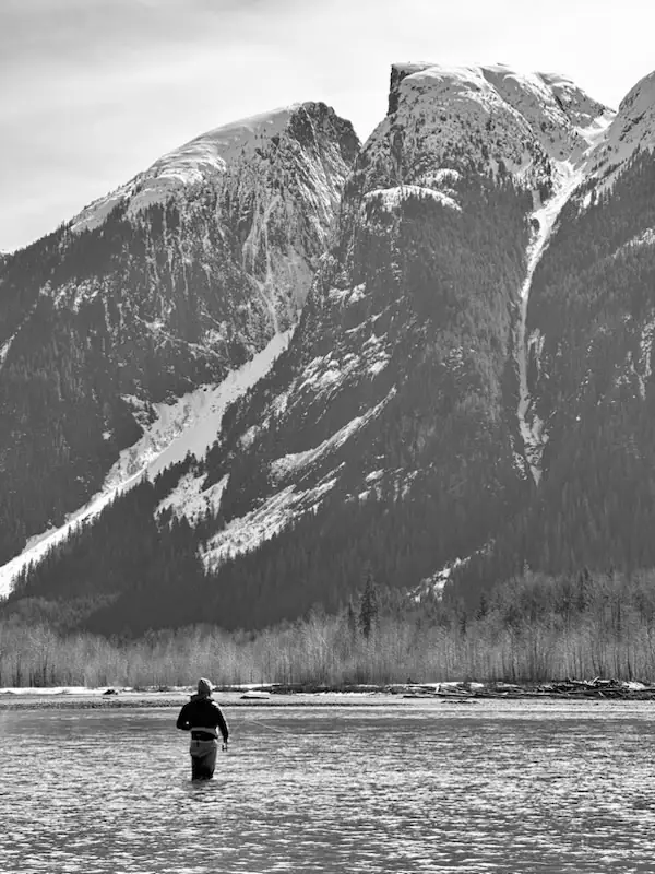 Kyle Ostenson wading a wide river beneath towering snow-covered mountains while steelhead fishing in a remote wild river valley.