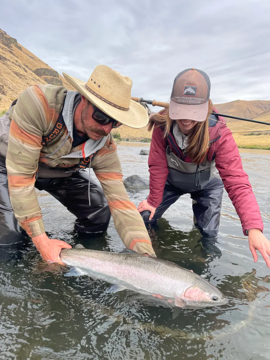 Nick Rowell steadying a large wild steelhead in the shallows of a high-desert Oregon river while a client leans in to help release the fish.