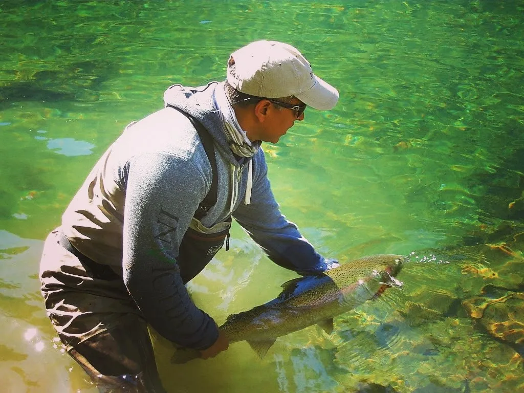 Pranjal “PJ” Srivastava standing in clear emerald shallows in waders and a ball cap, both hands supporting a heavily spotted wild rainbow trout as he eases it back into the current.