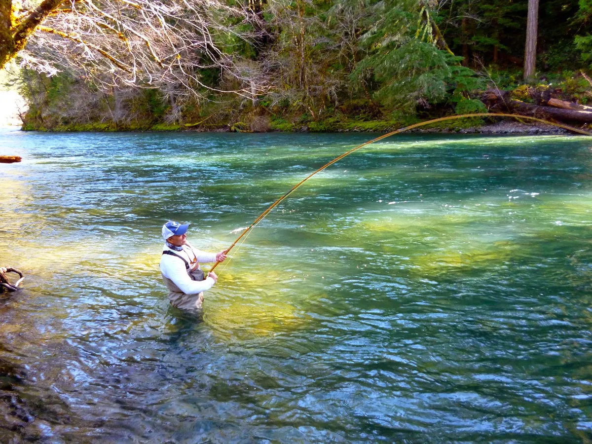 Pranjal “PJ” Srivastava standing waist-deep in a wide blue-green river below a forested bank, his two-handed rod bent deep into a running fish.