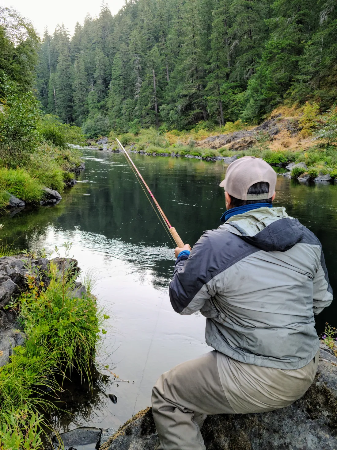 Pranjal “PJ” Srivastava seen from behind, seated on a mossy boulder with a long rod extended over a deep green pool framed by old-growth conifers on Oregon's North Umpqua River.