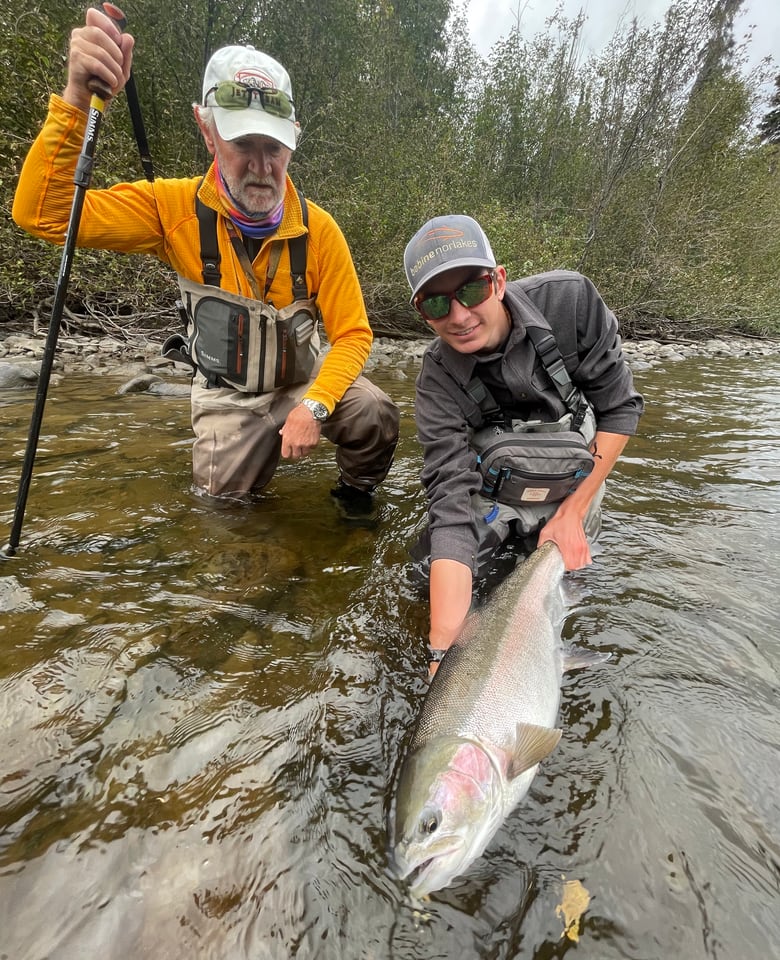 Logan Labonte kneeling in the shallows of the Babine River, both hands supporting a large chrome wild steelhead, with his grandfather beside him steadying a wading staff.